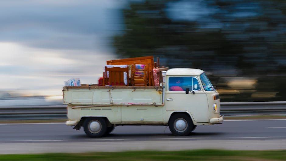 A vintage white van with a flatbed cargo area is in motion on a highway during daylight, with a blurred background indicating speed. The flatbed is loaded with household furniture and packing material