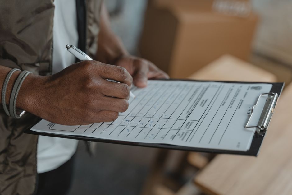 A person with dark skin, wearing a brown jacket and a grey wristband, is holding a pen and writing on a black clipboard that contains a moving or packing checklist with multiple boxes and rows of text